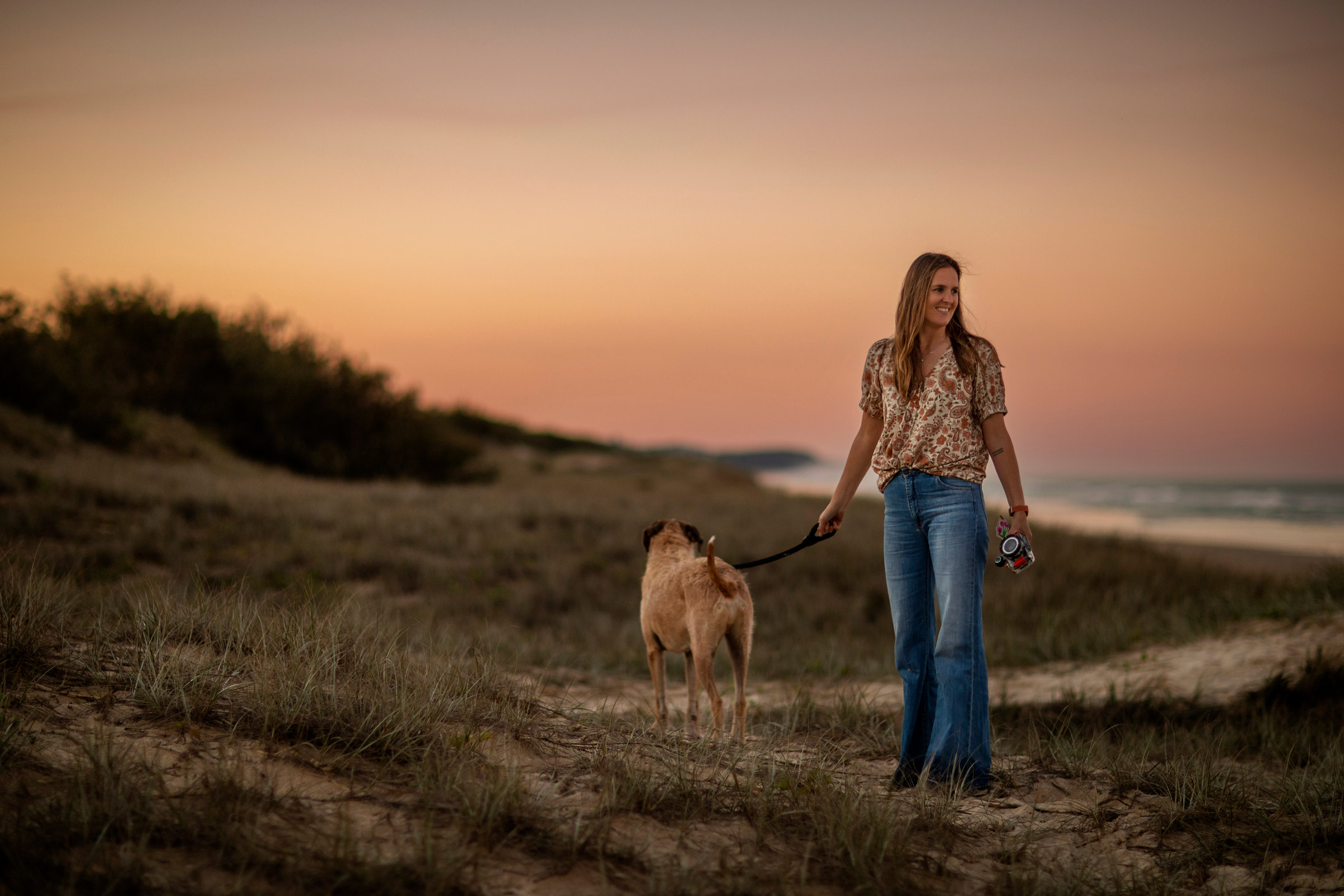 Woman walking her dog through coastal dunes at sunset